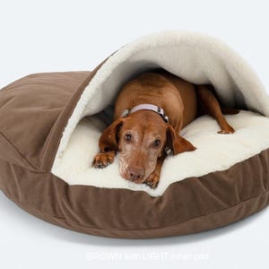 May include: A brown dog bed with a hooded design. The bed has a brown exterior and a soft, cream-colored interior. A brown dog with a pink collar is resting inside the bed.