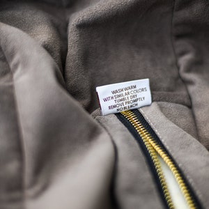 May include: Close-up of a gray suede jacket with a black zipper and a gold-colored zipper pull. A white tag with care instructions is visible. The tag reads: Wash warm with similar colors, tumble dry, remove promptly, no bleach.