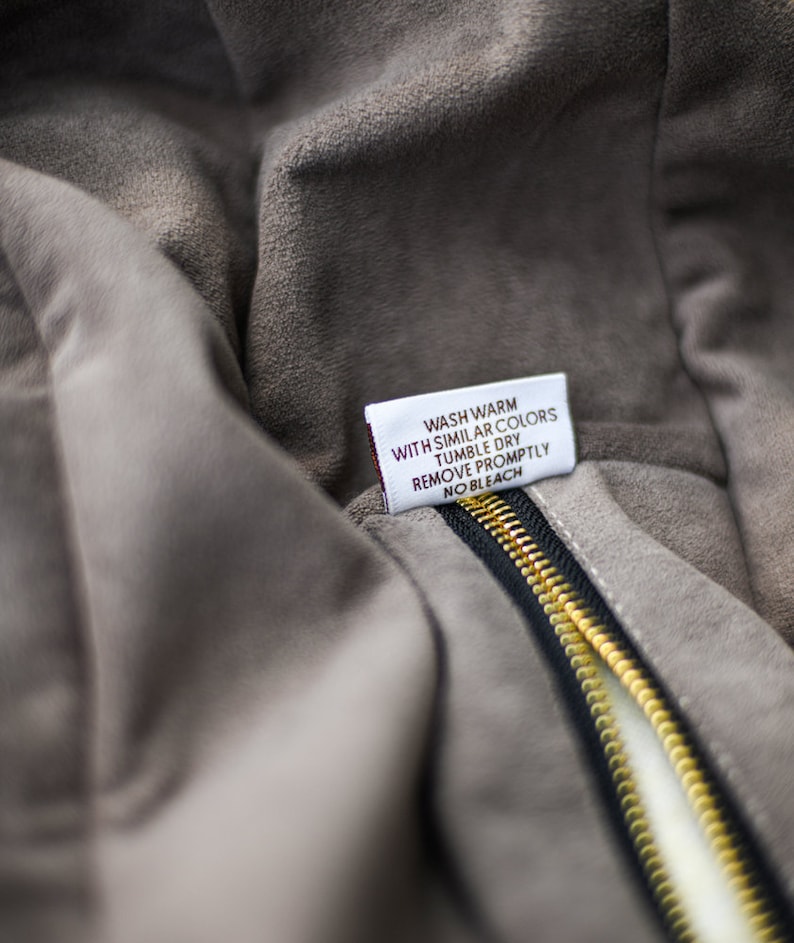 May include: Close-up of a gray suede jacket with a black zipper and a gold-colored zipper pull. A white tag with care instructions is visible. The tag reads: Wash warm with similar colors, tumble dry, remove promptly, no bleach.