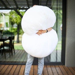 May include: A person holding two large, white, round objects, possibly pillows or cushions. The person is wearing blue jeans and standing barefoot on a wooden deck. The background includes a glass door and a glimpse of outdoor scenery.