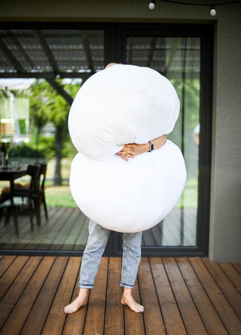 May include: A person holding two large, white, round objects, possibly pillows or cushions. The person is wearing blue jeans and standing barefoot on a wooden deck. The background includes a glass door and a glimpse of outdoor scenery.