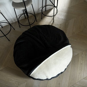 May include: A round, two-toned beanbag chair. The top half is black, and the bottom half is off-white. The chair is on a light wood floor. Three bar stools with black frames and white seats are in the background.
