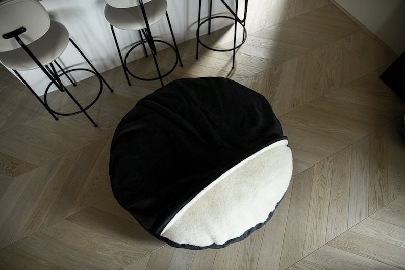 May include: A round, two-toned beanbag chair. The top half is black, and the bottom half is off-white. The chair is on a light wood floor. Three bar stools with black frames and white seats are in the background.