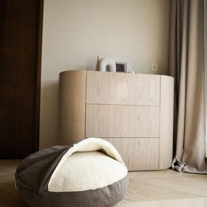 May include: A gray and white pet bed with a rounded shape, set on a light wood floor. The bed has a soft, plush interior and a dark gray exterior. A wooden dresser and a curtain are in the background.
