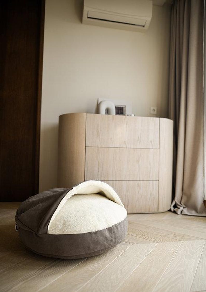 May include: A gray and white pet bed with a rounded shape, set on a light wood floor. The bed has a soft, plush interior and a dark gray exterior. A wooden dresser and a curtain are in the background.