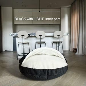 May include: A black and white pet bed with the text "BLACK with LIGHT inner part". The bed has a round shape with a black exterior and a white, cushioned interior. The bed is in a modern room setting.