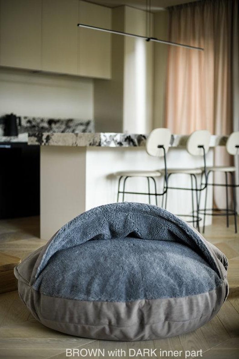 May include: A brown pet bed with a dark blue, plush interior. The bed has a rounded shape with a low profile and a raised, hooded section. The text "BROWN with DARK inner part" is visible at the bottom.