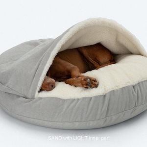May include: A light gray dog bed with a hooded top and a soft, cream-colored interior. A brown dog is curled up inside the bed, appearing asleep. The bed has a rounded shape and is made of a soft, plush material. The text "SAND with LIGHT inner part" is visible.