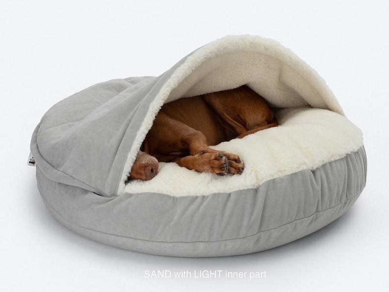 May include: A light gray dog bed with a hooded top and a soft, cream-colored interior. A brown dog is curled up inside the bed, appearing asleep. The bed has a rounded shape and is made of a soft, plush material. The text "SAND with LIGHT inner part" is visible.