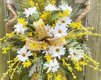 May include: A rustic wooden basket filled with a floral arrangement of white daisies and yellow flowers. The basket is decorated with a burlap bow with a yellow daisy print.