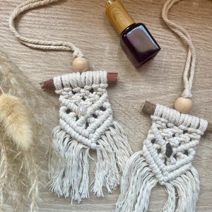Puede incluir: Dos ambientadores de coche de macramé color crema con detalles de madera y un pequeño frasco de aceite esencial morado oscuro con tapón de madera. Los ambientadores tienen flecos y cuentas de madera. Sobre una superficie de madera clara.