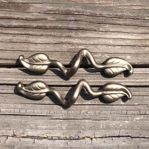 May include: Two antique bronze leaf-shaped drawer pulls. Each pull features a stylized leaf design at each end, connected by a curved, wave-like central element. The hardware is set against a weathered wooden surface.