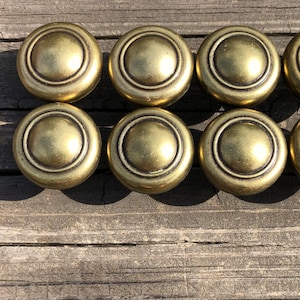 May include: Eight antique brass cabinet knobs with a round, stepped design. The knobs have a warm, golden hue and a slightly aged appearance, suggesting vintage or retro style. They are arranged in two rows on a weathered wooden surface.