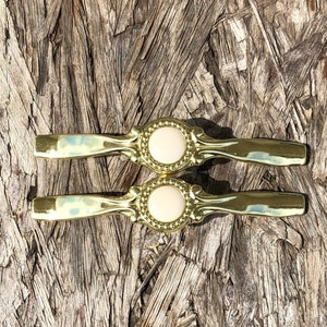 May include: Two gold cabinet knobs with a decorative design and a white center. The knobs are on a wood background.