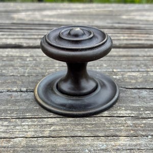 May include: A dark bronze-toned metal cabinet knob featuring a circular, tiered top design with concentric rings and a smooth, flared base. The knob has a matte finish and a central raised point, showcasing a classic, rounded silhouette for furniture.
