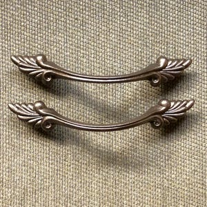 May include: Two antique bronze-colored drawer pulls. Each pull has a curved handle with decorative leaf-like accents on each end. The pulls are set against a textured, neutral-toned background.
