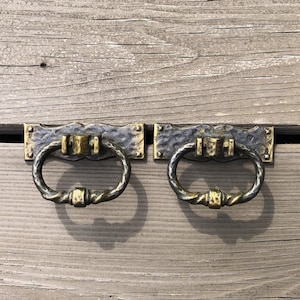 May include: Two antique-style drawer pulls with a black and gold finish. The pulls have a ring shape and are mounted on a rectangular plate.