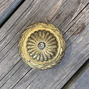 Op de afbeelding: Een ronde, antieke messing deurknop met een bloemenontwerp. De knop heeft een centraal bloemachtig patroon, omgeven door een gedetailleerde, getextureerde rand. Geplaatst op een verweerd houten oppervlak.