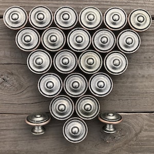 May include: A collection of 19 silver-toned cabinet knobs with a raised, circular design. The knobs are arranged in a heart shape on a wooden surface.