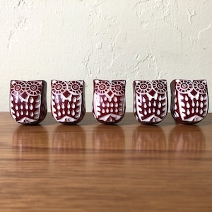 May include: Five maroon-colored owl figurines with white carved details. The owl figures are lined up on a wooden surface. Each owl has a detailed face and body design, with a symmetrical pattern.