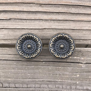 May include: Two antique bronze cabinet knobs with a floral design. Each knob features a central starburst pattern surrounded by a ring of small dots and a decorative outer edge. The knobs are set against a weathered wooden surface.