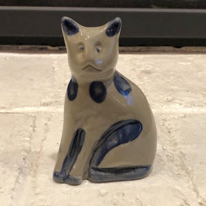 May include: A ceramic cat figurine in a seated position. The cat is primarily beige with dark blue accents, including spots and curved lines. The figurine has pointed ears and a slightly sad expression.