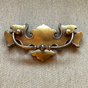 May include: A brass cabinet pull with a decorative, ornate design. The pull has a curved handle and two decorative knobs on either side.