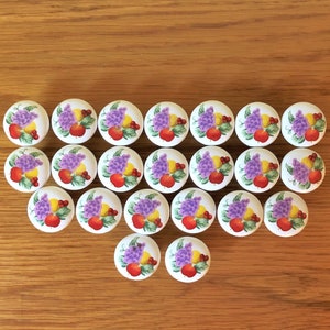 May include: A set of 18 round, white ceramic knobs with colorful fruit and floral designs. The knobs feature a variety of fruits, including grapes, apples, and lemons, as well as purple flowers. The knobs are arranged in a symmetrical pattern.