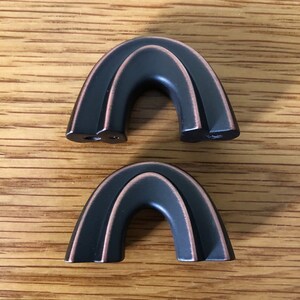 May include: Two arch-shaped cabinet pulls with a dark bronze finish and copper-toned highlights along the edges. Each piece features a smooth, curved design with two mounting points visible on the underside of the top pull and a solid base.