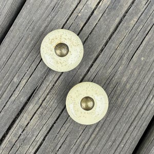 May include: Two round cabinet knobs featuring a glossy cream-colored ceramic surface with fine brown speckles. Each knob is centered with a smooth, circular metallic brass-toned accent piece that sits slightly raised from the main body.