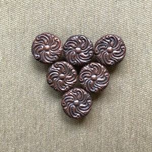 May include: Six brown metal buttons with a decorative floral design arranged in a triangular pattern on a beige fabric background.