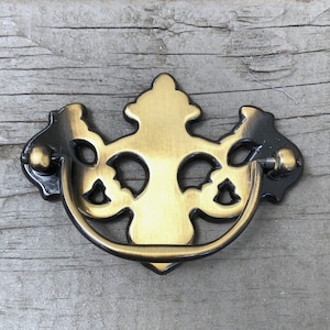May include: A decorative drawer pull with a brass finish and black accents. The pull features an ornate design with a curved handle and decorative backplate. The hardware is set against a weathered, gray wooden surface.