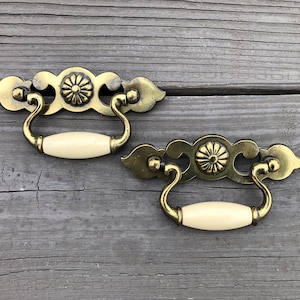 May include: Two antique brass cabinet pulls with ornate floral designs and off-white ceramic handles.
