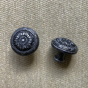May include: Two decorative cabinet knobs with an antique silver finish. The knobs have a detailed, embossed floral design. One knob is a round pull, and the other has a short stem for mounting. The knobs are on a textured, tan surface.