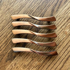 May include: Five cabinet pulls arranged in a vertical stack. Each pull features a curved, twisted design with a brushed copper finish that transitions into darker, metallic tones at the ends, creating a distinct gradient effect across the surface.