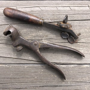 May include: Two antique metal tools with wooden handles rest on a weathered wooden surface. One tool has a curved handle and a jaw-like structure, while the other has a wooden handle and a metal head with adjustable components. The tools appear to be vintage.