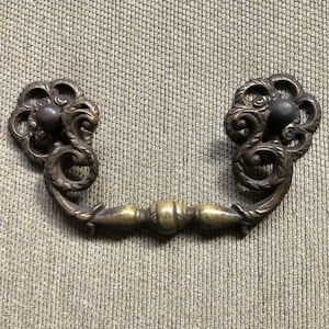 May include: A brass drawer pull with ornate scrollwork and a round knob on each end. The pull is in a U-shape and has a decorative center piece.