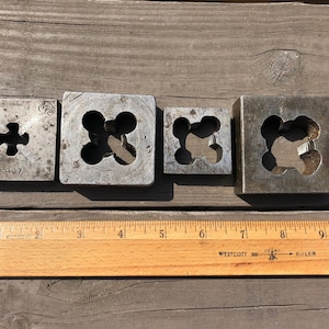 May include: Four metal square dies with different sized holes in the center. The dies are arranged in a row from left to right, with the smallest die on the left and the largest die on the right. The dies are all made of a dark gray metal and have a rough texture. The dies are sitting on a wooden surface. The text "PAT. JAN. 9-07" is visible on the smallest die.