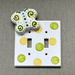 May include: A white double light switch plate featuring a raised ceramic butterfly accent in yellow and green. The plate is decorated with a pattern of yellow and light green polka dots and includes two rectangular cutouts for toggle switches.