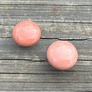 May include: Two smooth, round, glossy cabinet knobs in a solid dusty peach color. Each knob features a spherical head with a reflective surface, designed for attachment to furniture or cabinetry doors and drawers.