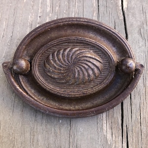 May include: A vintage, oval-shaped, cast iron cabinet pull with a decorative, swirling design in the center. The pull is a dark brown color and has a slightly distressed finish.