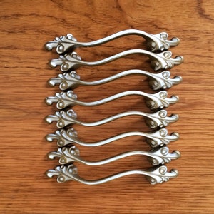 May include: Eight silver-toned cabinet pulls with a decorative scroll design. The pulls are arranged in a row, with the top pull at the top of the image and the bottom pull at the bottom of the image.