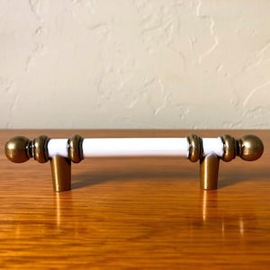May include: White cabinet knob with brass accents. The knob has a cylindrical shape with rounded ends and two brass rings on each side.