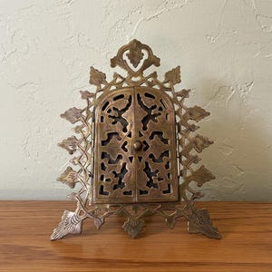 May include: A brass-colored metal frame featuring two hinged doors with intricate cut-out patterns. The frame has a pointed arch shape with decorative leaf-like protrusions extending from the edges and a small central knob on the doors.