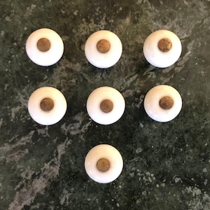 May include: Seven white cabinet knobs with a brass-colored center are arranged on a dark green marble surface. The knobs are round with a slightly domed shape, and the brass center adds a touch of contrast. These knobs are suitable for furniture restoration or new builds.