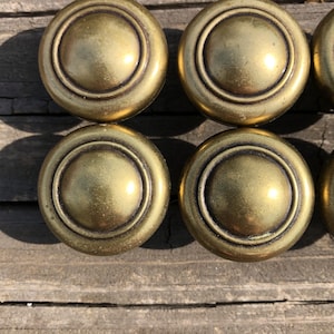 May include: A set of four round brass cabinet knobs featuring a smooth, domed center and a raised circular rim. The metal has a warm, aged gold finish with subtle dark patina accents around the edges, creating a consistent look across all pieces.