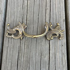May include: A brass-toned metal drawer pull featuring an ornate, symmetrical design with curved, scroll-like flourishes on both ends and a central handle bar. The hardware has a weathered, antique finish with visible dark patina in the crevices.