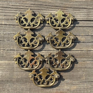 May include: A collection of seven matching brass drawer pulls, each featuring an ornate, symmetrical openwork backplate with a curved, swinging bail handle. The metal has a warm, aged golden patina with visible surface texture and detailing.