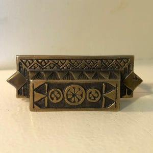 May include: A bronze-colored drawer pull with geometric and floral designs. The rectangular pull features a decorative top with a repeating pattern, a central section with floral motifs, and diamond-shaped accents on each end. The hardware is likely for a cabinet or drawer.