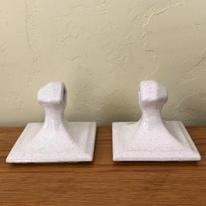 May include: Two white ceramic towel holders with a speckled design. Each holder has a square base and a curved handle. The holders are displayed on a wooden surface against a light-colored wall.